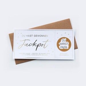 May include: A white rectangular lottery ticket with gold foil accents and the German text DU HAST GEWONNEN Jackpot, featuring a circular gold scratch-off area with the hidden text DU WIRST PAPA, placed on top of a brown paper envelope.