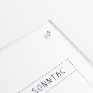 May include: A clear acrylic calendar with the word "SONNTAG" printed on it. The calendar has a hole in the top corner for hanging.