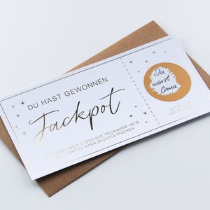 May include: A white rectangular scratch-off lottery ticket with gold foil accents and stars. The text reads DU HAST GEWONNEN Jackpot, with a gold circle revealing the handwritten text Du wirst Oma. The ticket rests on a brown paper envelope.