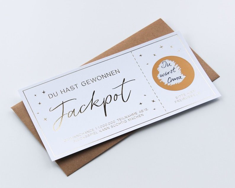 May include: A white rectangular scratch-off lottery ticket with gold foil accents and stars. The text reads DU HAST GEWONNEN Jackpot, with a gold circle revealing the handwritten text Du wirst Oma. The ticket rests on a brown paper envelope.