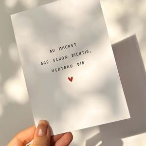 Può includere: Una carta bianca con testo scritto a mano nero e un piccolo cuore rosso. Il testo recita "DU MACHST DAS SCHON RICHTIG, VERTRAU DIR". La carta è tenuta su uno sfondo bianco con ombre.