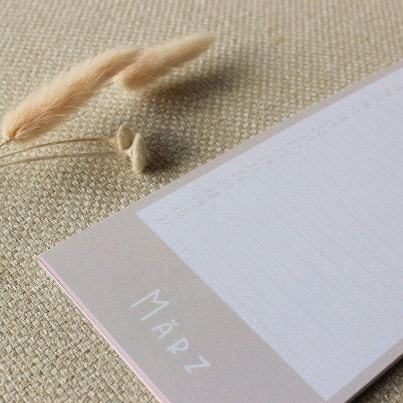 May include: A beige monthly calendar notepad with the word "M&Auml;RZ" printed on the front. The notepad has lined pages for writing notes and appointments. The notepad is lying on a beige textured surface.