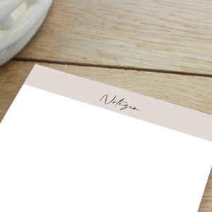 Puede incluir: Un bloc de notas blanco con un encabezado beige que dice "Notizen" en cursiva negra.