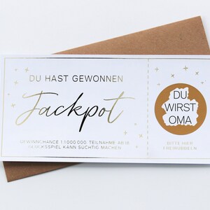 May include: A white rectangular scratch-off lottery ticket with gold foil accents and text that reads DU HAST GEWONNEN Jackpot, with a circular gold scratch-off area containing the text DU WIRST OMA, placed over a brown paper envelope.