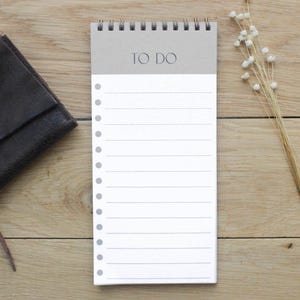 Może przedstawiać: Biały notes z brązową okładką i spiralnym oprawą. Na okładce widnieje napis "TO DO". Notes leży na drewnianej powierzchni.