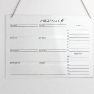 May include: A clear acrylic weekly planner with a black and white design. The planner includes sections for each day of the week, a to-do list, and a meal plan. The text "UNSERE WOCHE" is printed at the top of the planner.