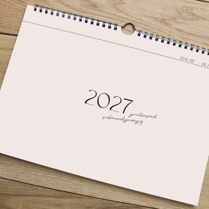 Może przedstawiać: Kalendarz ścienny z jasnor&oacute;żową okładką, na kt&oacute;rej widnieje rok 2027 dużą czarną czcionką szeryfową, z niemieckim napisem "zweitausend siebenundzwanzig" poniżej oraz "JANUAR - DEZEMBER" w prawym g&oacute;rnym rogu, oprawiony czarną spiralą.
