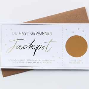 May include: A white rectangular scratch-off ticket with gold foil accents and text that reads DU HAST GEWONNEN Jackpot, featuring a circular gold scratch area on the right side with the text BITTE HIER FREIRUBBELN, resting on a brown envelope.