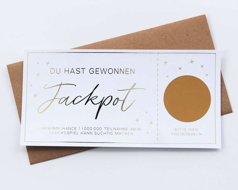 May include: A white rectangular scratch-off ticket with gold foil accents and text that reads DU HAST GEWONNEN Jackpot, featuring a circular gold scratch area on the right side with the text BITTE HIER FREIRUBBELN, resting on a brown envelope.