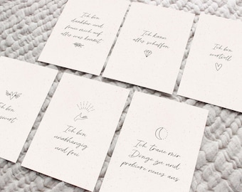 May include: A set of cream-coloured affirmation cards with black handwritten text and small illustrations. The cards feature phrases like "I am grateful" and "I can do anything." The cards are arranged on a textured, light-coloured surface.
