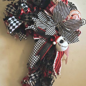 May include: A decorative candy cane-shaped wreath with a Halloween theme. It features black and white striped, checkered, and houndstooth ribbons, along with red accents. A Jack Skellington face ornament is attached, and the wreath is adorned with spiderweb and other spooky embellishments.