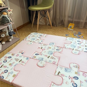 Puede incluir: Una alfombra grande compuesta por piezas de espuma entrelazadas en forma de rompecabezas. La alfombra presenta secciones alternas de tela rosa claro lisa con pequeños puntos blancos y tela blanca estampada con ilustraciones de niñas, bicicletas y juguetes.