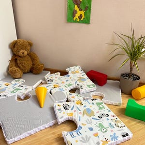 May include: Large foam puzzle floor mats featuring a white surface with colorful illustrations of various animals including elephants, monkeys, and giraffes, paired with gray textured mats. The edges are finished with a white dimpled fabric trim.