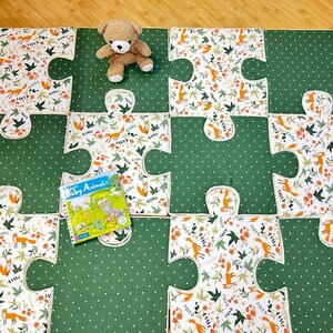 May include: A set of interlocking floor mats shaped like puzzle pieces, featuring alternating patterns of white fabric with orange foxes and green fabric with white polka dots. A small brown plush teddy bear and a book titled Baby Animals sit on top.