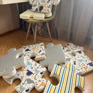 May include: A collection of puzzle-shaped floor cushions featuring various fabric patterns, including a white background with colourful dinosaur illustrations, a grey and white chevron print, and a blue, yellow, and white vertical stripe design.