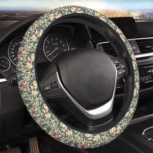 May include: Steering wheel cover featuring a cream-coloured background with a floral pattern in red, blue, and green. The cover is fitted on a black steering wheel inside a car.