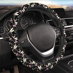 May include: A black steering wheel cover with a floral pattern featuring pink, white, and yellow flowers and green leaves. The cover is installed on a car's steering wheel, with the dashboard and other interior details visible in the background.