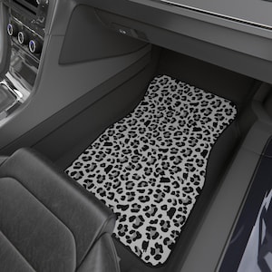 May include: A car floor mat with a black, white, and gray leopard print pattern. The mat is rectangular with rounded edges and is placed inside a car. The interior of the car is black and gray.