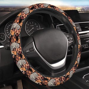 Pode incluir: Uma capa de volante preta com um padrão repetido de várias raças de cães em tons de castanho, cinza e creme sobre um fundo preto. A capa está instalada no volante de um carro, com o painel e outros elementos interiores visíveis.