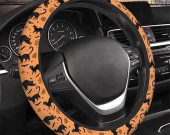Könnte beinhalten: Ein Halloween-Lenkradbezug mit einem sich wiederholenden Muster aus schwarzen Katzen, Fledermäusen und Monden auf orangefarbenem Hintergrund. Der Bezug ist an einem Lenkrad eines Autos angebracht, wobei das Armaturenbrett und andere Innenelemente sichtbar sind.