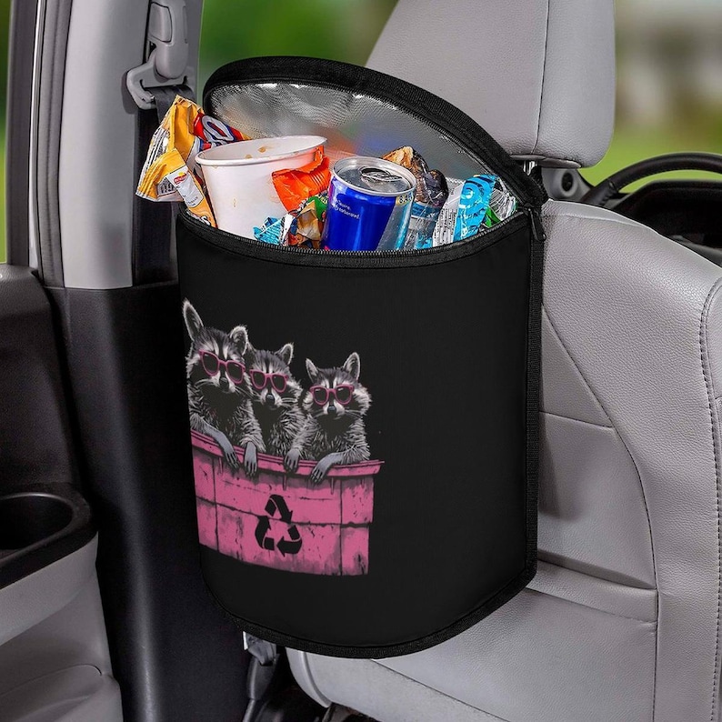 May include: Black car trash bag with a funny raccoon design. The bag is hanging on a car seat. The bag has a black exterior with a graphic of three raccoons wearing sunglasses in a pink trash bin. The bin has a pink recycle symbol. The interior is silver and insulated. The bag is open and filled with trash.