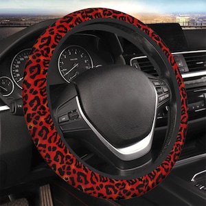 Pode incluir: Uma capa de volante com um design de estampa de leopardo vermelho. A capa está enrolada em um volante preto, com o painel e o interior do carro visíveis ao fundo. Um acessório decorativo.