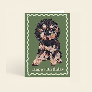 Könnte beinhalten: Geburtstagskarte mit einer Aquarellillustration eines kleinen Hundes mit schwarzem und braunem Fell. Der Hund ist vor einem grünen Hintergrund mit einem weißen, wellenförmigen Rand platziert. Die Worte "Happy Birthday" sind unten aufgedruckt.