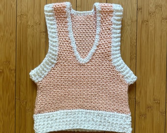 May include: A sleeveless crochet vest featuring a peach-colored main body with a V-neckline. The garment is accented with thick, white ribbed trim along the armholes, the neckline, and the bottom hem, creating a distinct contrast in color and texture.