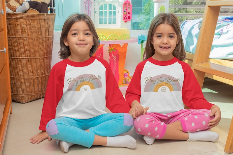 May include: Two children wearing red and white baseball shirts with a rainbow and the text "Play Time Blanks" printed on the front. The children are sitting on the floor with their legs crossed.