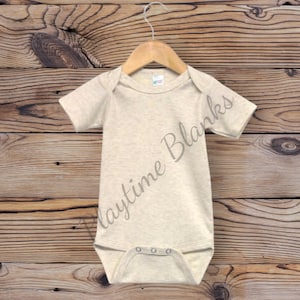 May include: A beige short-sleeved baby onesie with snaps at the bottom. The onesie is hanging on a wooden hanger against a woodgrain background.