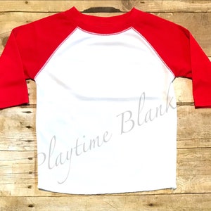 May include: White long-sleeve baseball t-shirt with red sleeves. The shirt has a white trim around the neck and sleeves.