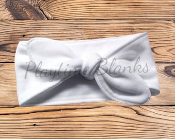 May include: White knotted headband made of soft fabric. The headband is tied in a bow at the top. The headband is on a wooden background.