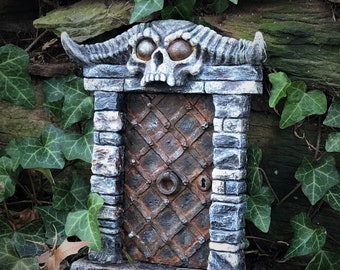 May include: A miniature stone door with a skull and horns above it. The door is made of metal and has a rusty finish. The door is surrounded by a stone frame and has a keyhole.
