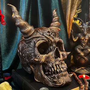 Puede incluir: Un cráneo de color marrón oscuro, tallado intrincadamente, con cuernos, se sienta sobre un libro negro con una moneda de oro en la parte superior. El cráneo está rodeado de velas, un collar de cuentas y una pequeña estatua de un demonio.