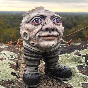 May include: A gray and brown ceramic figurine of a person with a large head and a moon-like face. The figurine has green eyes, a pink mouth, and a gray and brown body with legs and feet.