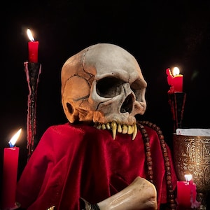 May include: A detailed skull with sharp teeth rests on a red cloth, surrounded by lit red candles. The skull has visible cracks and a detailed jawline. The scene is set against a dark background, creating a gothic or Halloween theme.