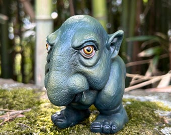 以下が含まれることがあります： 大きく垂れ下がった鼻と大きな黄色い目をした生き物の小さな緑色の手描き人形。生き物は質感のある表面をしていて、粘土でできているように見える。