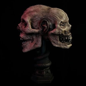 May include: A two-faced skull sculpture with a weathered, textured appearance. The sculpture is primarily tan and brown, with hints of red. It is mounted on a black stand with a snake-like design, set against a black background.