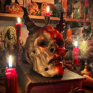 May include: A detailed, horned skull mask with a weathered, textured appearance, stained with red accents. The mask rests on a thick, antique book, surrounded by lit red candles and other occult-themed objects.