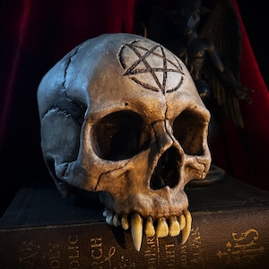 Puede incluir: Una calavera detallada y desgastada con dientes prominentes y un pentagrama grabado en la frente. La calavera tiene una apariencia realista y envejecida, con grietas e imperfecciones visibles. La estética general sugiere un tema gótico u oculto.