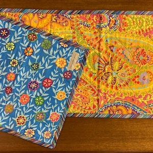 May include: Two quilted table runners with floral patterns. One runner is blue with white and multi-colored flowers, the other is yellow with pink, orange, and purple flowers.