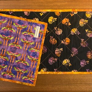 May include: A reversible quilted table runner with an orange border. One side features a purple fabric with a repeating green and orange abstract pattern. The other side has a black fabric with a repeating orange pumpkin print and a 24-inch ruler.