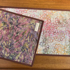 May include: A rectangular quilted table runner featuring two distinct fabric patterns with a brown border. One side displays a purple and green abstract leaf print, while the other shows a floral pattern in pink, orange, and white. A small white label is attached.