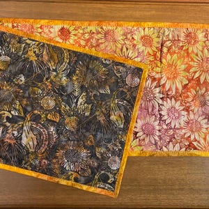 May include: A rectangular quilted table runner featuring a reversible design with a sunflower pattern. One side displays dark gray and black sunflowers, while the other side shows orange and yellow sunflowers. A wooden ruler is visible on the right.