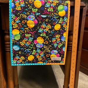 May include: A quilted table runner with a black background, featuring colorful balloons, ribbons, and the words "Happy Birthday." The runner has a teal border with yellow trim and a diamond stitch pattern.