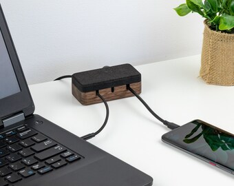 May include: A wooden cable organizer with a black felt top and three slots for cords. A laptop and a smartphone are plugged into the organizer, which is sitting on a white desk.