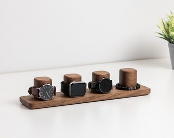 May include: A wooden watch display stand with four watch holders. The stand holds four watches with different bands and faces. A small potted plant sits in the background. The stand is made of dark wood and sits on a white surface.