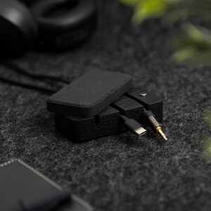 May include: A black wooden box with a hinged lid, designed to store and organize cables and cords. The box is open, revealing a black cable and a gold-tipped audio cable.