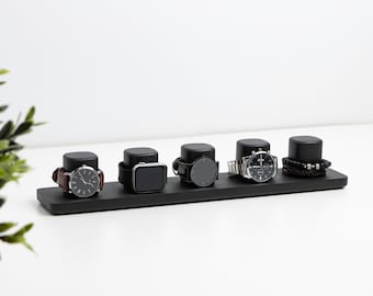 Op de afbeelding: Een zwarte horlogehouder met zes vakken voor horloges en armbanden. De houder is gemaakt van hout en heeft een strak, modern ontwerp. Hij heeft vijf horloges en één armband.