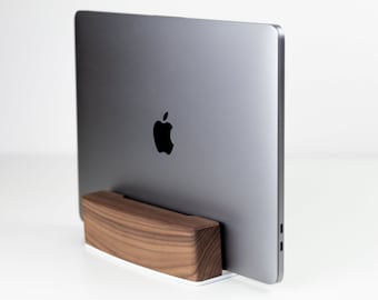 May include: A silver laptop computer is sitting on a wooden stand. The stand is made of dark wood with a natural grain pattern. The laptop is closed and the Apple logo is visible on the back.